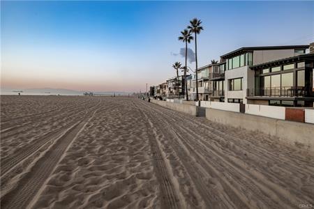Empty sandy beach at sunset with a row of modern houses and palm trees along the boardwalk; tire tracks crisscross the foreground.
