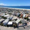 Aerial view of a coastal town with dense grid of houses along a curved street, ocean beyond the beach under a clear blue sky.