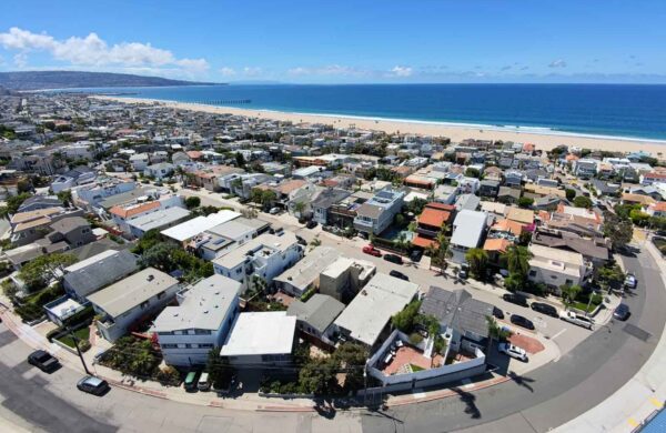 Aerial view of a coastal town with dense grid of houses along a curved street, ocean beyond the beach under a clear blue sky.